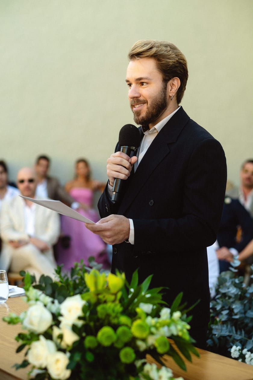 Rafael sorrindo, com o microfone e o roteiro na mão, conduzindo uma cerimônia de casamento